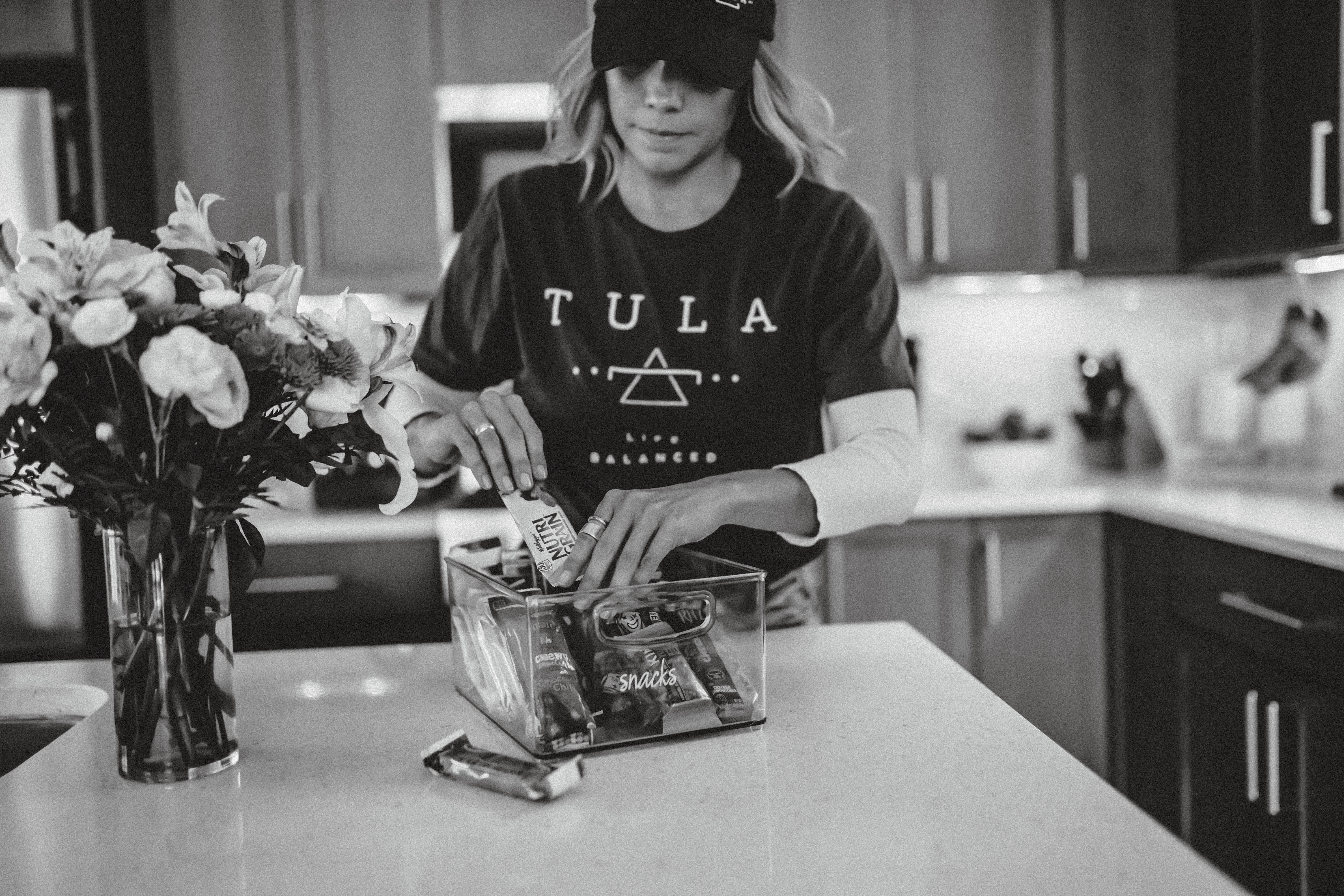 TULA Assistant wears TULA shirt and hat as she organizes a snack clear container on a white countertop next to a vase of flowers